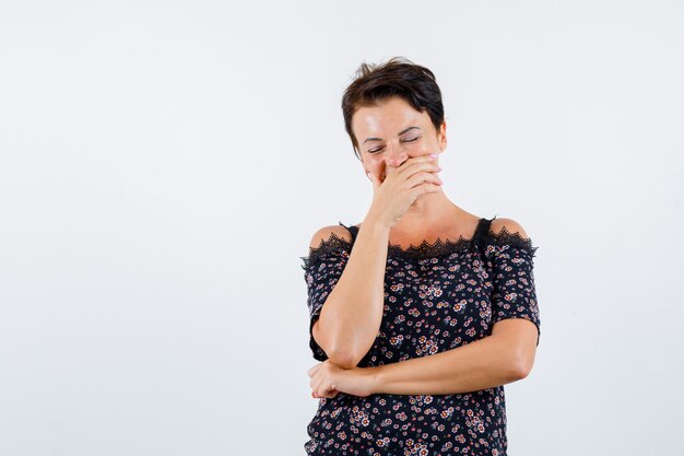 Reife Frau in Blumenbluse, schwarzer Rock, der Mund mit Hand bedeckt, lachend und fröhlich aussehend, Vorderansicht.