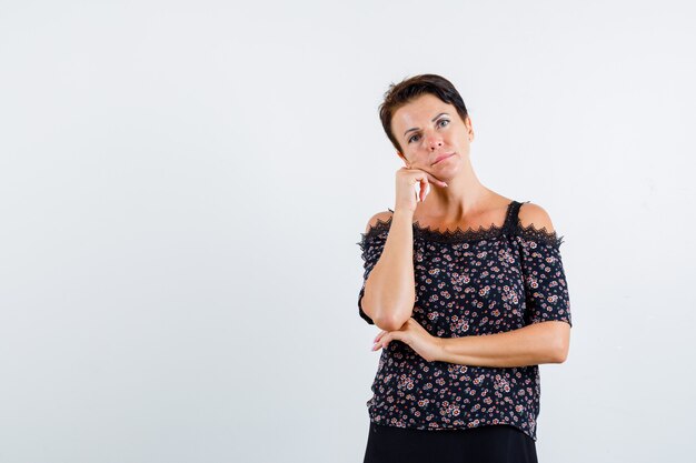 Reife Frau in Blumenbluse und schwarzem Rock lehnt Kinn an Hand, hält eine Hand unter Ellbogen und sieht nachdenklich aus, Vorderansicht.