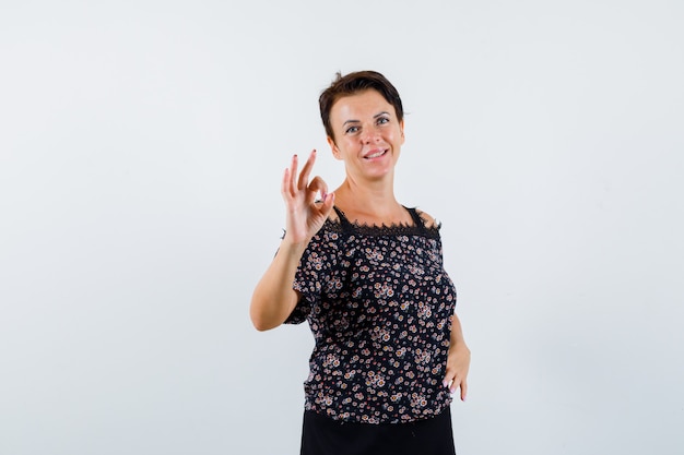 Reife Frau in Blumenbluse und schwarzem Rock zeigt ok Zeichen und schaut fröhlich, Vorderansicht.