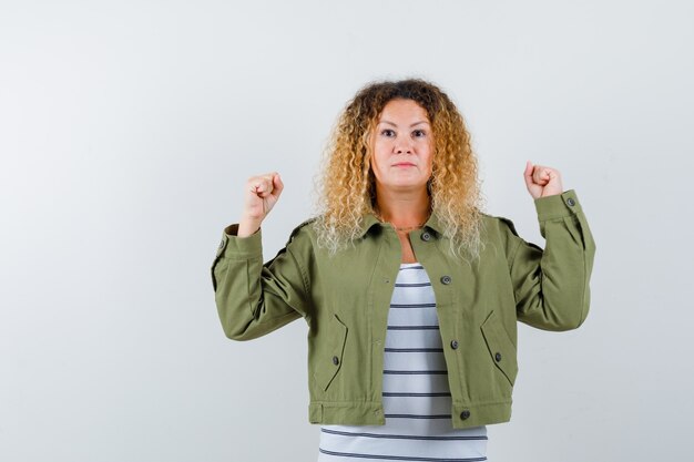 Reife Frau in grüner Jacke, T-Shirt zeigt Siegergeste und siegreich aussehend, Vorderansicht.