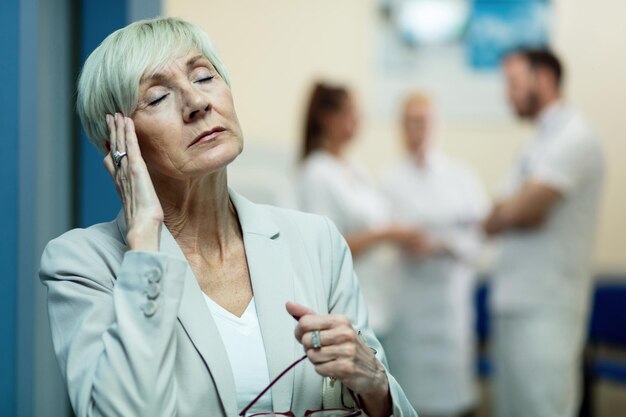 Reife Frau mit geschlossenen Augen, die ihren Kopf vor Schmerzen hält, während sie im Krankenhausflur Kopfschmerzen hat