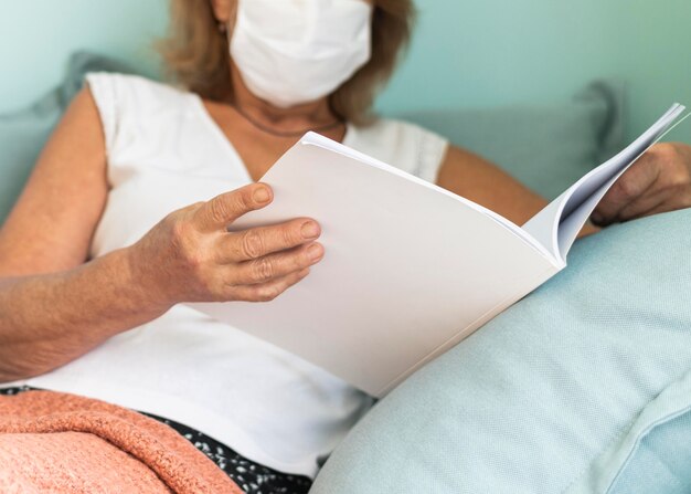 Reife Frau mit medizinischer Maske zu Hause während der Pandemie, die ein Buch liest