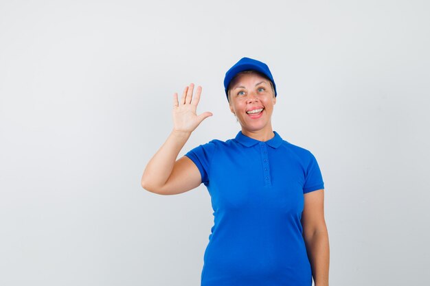 Reife Frau winkt Hand zum Begrüßen im blauen T-Shirt und schaut froh.