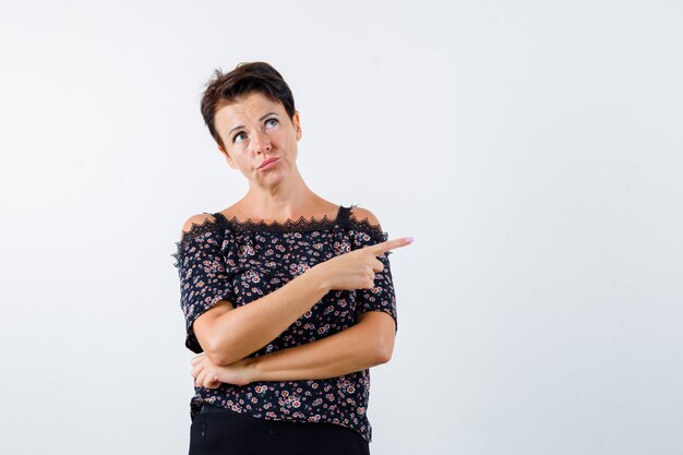 Reife Frau zeigt auf die rechte Seite, während sie Lippen in Bluse schmollt und nachdenklich aussieht. Vorderansicht.