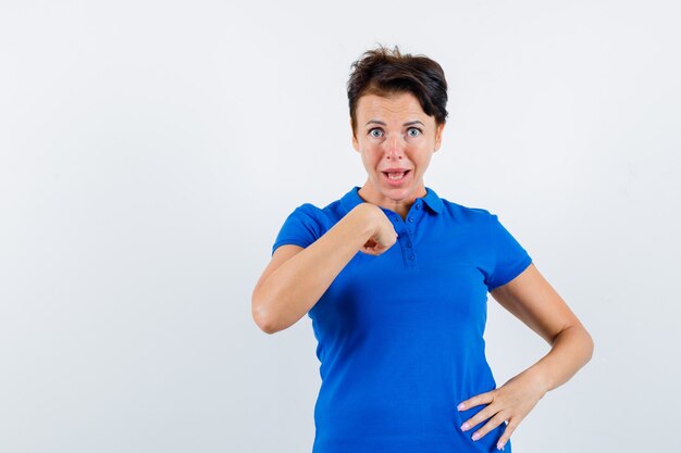 Reife Frau zeigt auf sich selbst in verwirrter Geste im blauen T-Shirt und schaut aufgeregt, Vorderansicht.