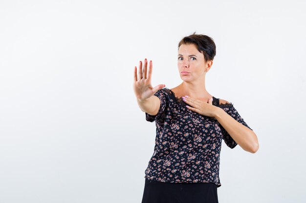 Reife Frau zeigt Stoppschild, hält Hand über Brust in Blumenbluse und schwarzem Rock und sieht ernst, Vorderansicht.