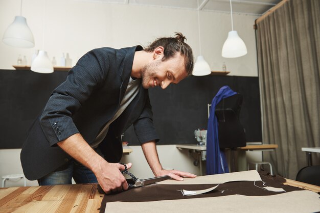 Reife freudige attraktive dunkelhaarige hispanische, männliche Modedesignerin, die in der Werkstatt an einem neuen Kleid arbeitet, Teile ausschneidet, Muster macht, Teile zusammennäht.