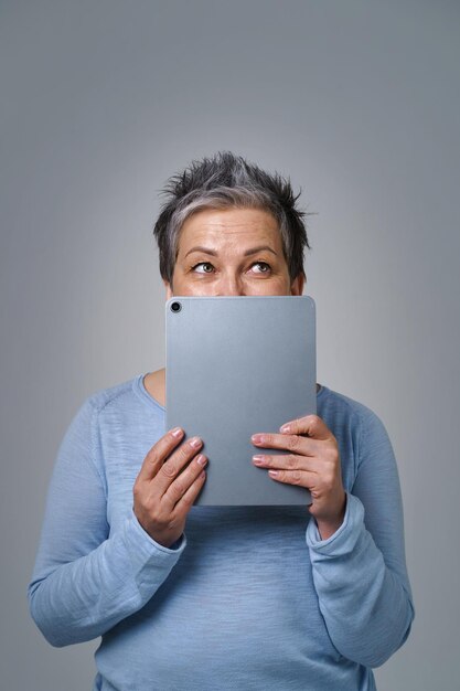 Reife grauhaarige Frau verstecken Gesicht schüchtern lächelnd hinter Smartphone oder modernem Gadget, das online arbeitet oder einkauft oder soziale Medien überprüft Hübsche Frau in blauer Bluse isoliert auf gelbem Hintergrund