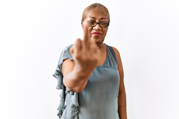 Reife hispanische Frau mit Brille, die vor isoliertem Hintergrund steht und Mittelfinger, unhöflichen und unhöflichen Gesichtsausdruck zeigt