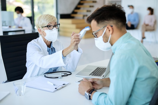 Reifer Arzt misst die Temperatur des Mannes mit Infrarot-Thermometer während des Termins in der medizinischen Klinik