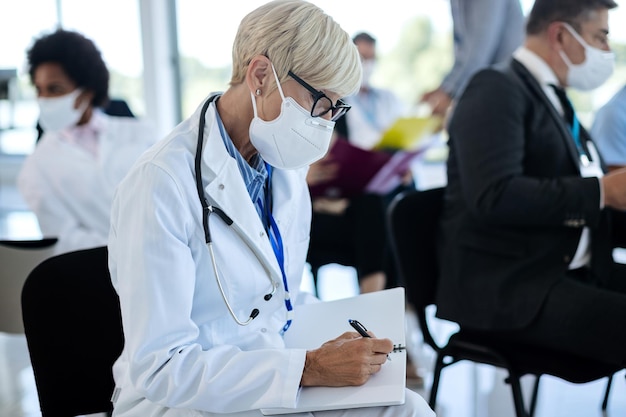Reifer Arzt mit Gesichtsmaske, der sich Notizen macht, während er an einer Bildungsveranstaltung im Konferenzsaal teilnimmt