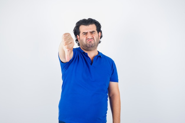 Reifer Mann, der Daumen unten im blauen T-Shirt, Jeans zeigt und enttäuscht schaut, Vorderansicht.