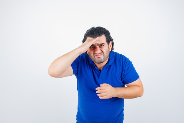 Reifer Mann, der weit weg mit Hand über Kopf in blauem T-Shirt, Jeans schaut und konzentriert schaut. Vorderansicht.