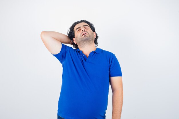 Reifer Mann im blauen T-Shirt, das Hand hinter Kopf hält und schläfrig sieht, Vorderansicht.