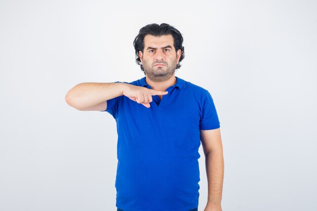 Reifer Mann im blauen T-Shirt, zeigt mit dem Zeigefinger nach rechts und sieht wütend aus, Vorderansicht.