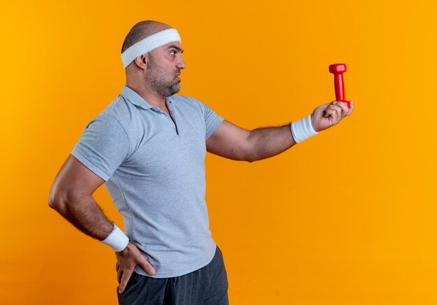 Reifer sportlicher Mann im Stirnband, der Hantel hält, der es mit ernstem Gesicht betrachtet, das über orange Wand steht