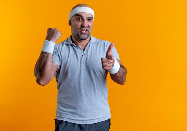 Reifer sportlicher Mann im Stirnband, der mit dem Finger auf die vordere geballte Faust zeigt, die zuversichtlich steht, über orange Wand zu stehen