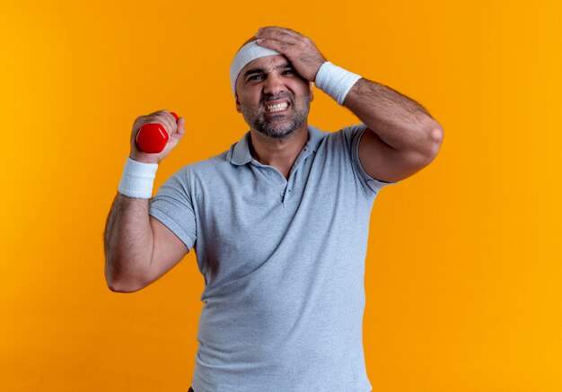 Reifer sportlicher Mann im Stirnband, der mit Hantel arbeitet, die verwirrt steht über orange Wand steht