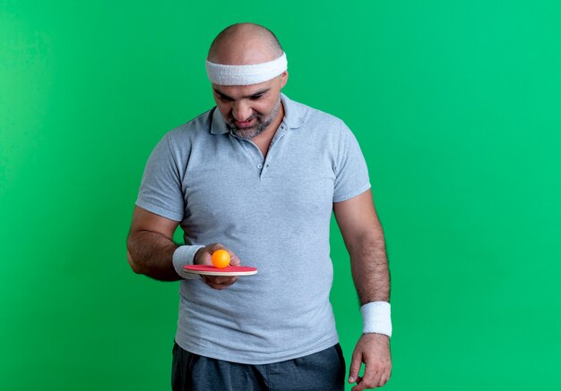 Reifer sportlicher Mann im Stirnband, der Schläger mit Ball für Tischtennis hält, der es mit Interesse betrachtet, das über grüner Wand steht