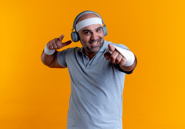 Reifer sportlicher Mann im Stirnband mit Kopfhörern auf seinem Kopf, der mit dem Finger nach vorne zeigt und fröhlich über orange Wand steht