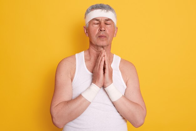 Reifer weißhaariger Mann, der weißes T-Shirt und Stirnband trägt, die Augen geschlossen hält, Hände zusammenhält und für ein besseres Leben betet
