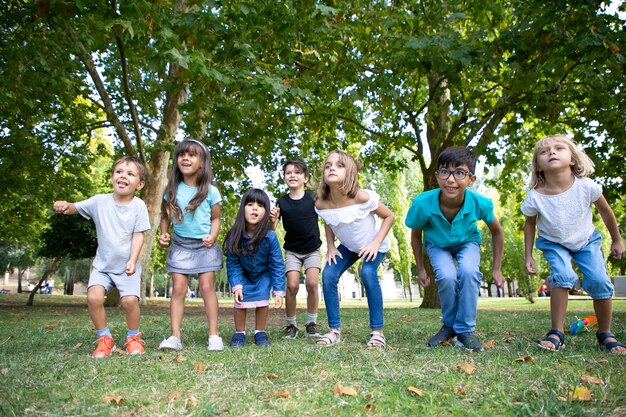 Reihe fröhlicher Kinder, die zusammen im Park Kniebeugen machen und vor Aufregung wegschauen. Kinderparty oder Unterhaltungskonzept