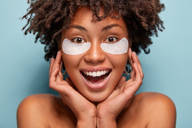 Reine schöne Frau kümmert sich um die Haut, hat unter Augenklappen, hält die Hände im Gesicht, lächelt breit, hat weiße Zähne, Afro-Haarschnitt, Modelle über blauer Wand. Schönheits- und Kosmetikkonzept