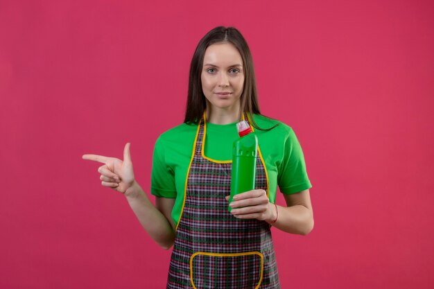 Reinigung der jungen Frau, die Uniform hält, die Reinigungsmittel hält, zeigt Finger zur Seite auf isolierte rosa Wand