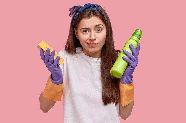 Reinigungs- und Reinigungskonzept. Unglückliche verwirrte junge Hausmeisterin mit langen Haaren, hält Schwamm und grüne Flasche mit Waschmittel, trägt Gummihandschuhe zum Schutz der Hände, erledigt Hausarbeiten