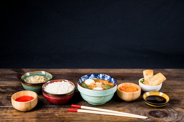 Reis; gekeimte Bohnen; Frühlingsrollen; Fischballsuppe und -soßen mit Stäbchen auf Tisch vor schwarzem Hintergrund