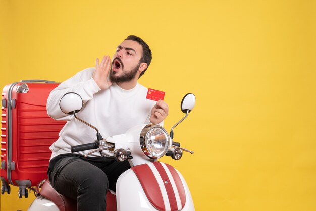 Reisekonzept mit einem jungen reisenden Mann, der auf einem Motorrad mit Koffer darauf sitzt und eine Bankkarte hält, die jemanden auf Gelb anruft