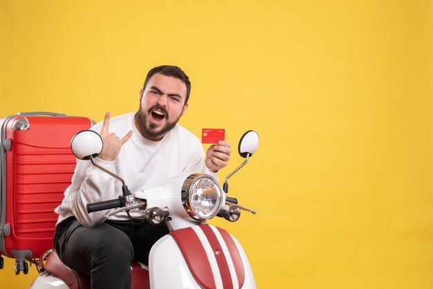 Reisekonzept mit jungen verrückten emotionalen lustigen reisenden Mann, der auf einem Motorrad mit Koffer darauf sitzt und Bankkarte auf Gelb hält holding