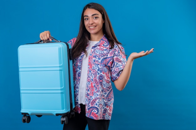reisende Frau, die mit Reisekoffer mit Lächeln auf Gesicht steht, das mit Arm der Hand auf lokalisiertem Blau präsentiert