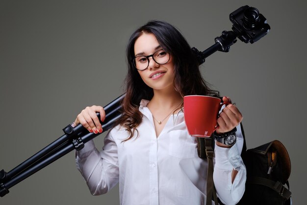 Reisende Frau hält digitale Fotokamera und eine rote Kaffeetasse über grauem Hintergrund.