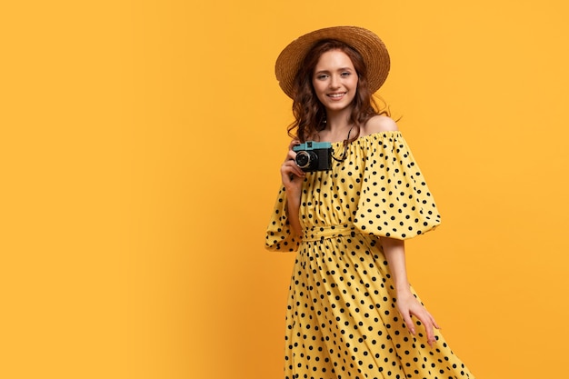 Reisende Frau in Strohhut und Sommerkleid posiert mit Retro-Kamera auf Gelb