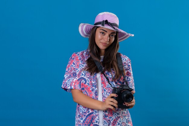 Reisende Frau mit Hut mit Fotokamera mit Lächeln auf Gesicht positiv und glücklich stehend auf isoliertem Blau