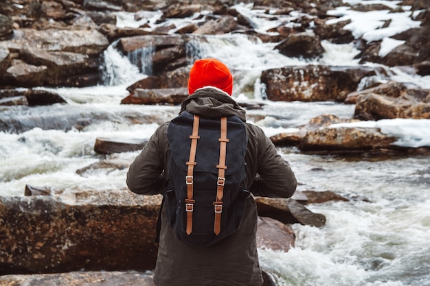 Reisender mann mit einem rucksack, der auf einem felsen steht, die hände auf der seite auf dem bergfluss und dem wasserfall