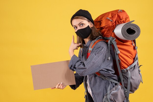 Reisendes Mädchen mit schwarzer Maske und Rucksack mit Pappe, die nach hinten zeigt