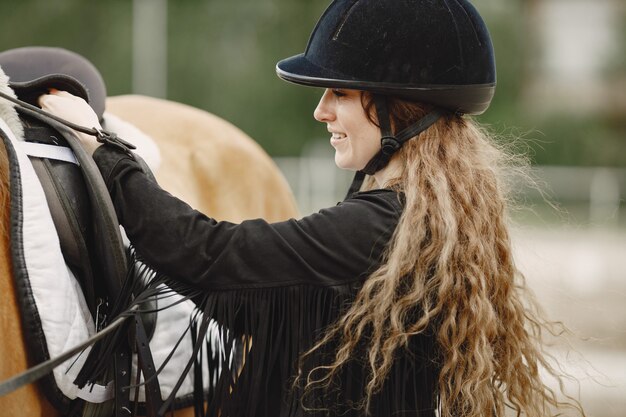 Reiterfrau, die mit ihrem Pferd auf einer Ranch spricht Frau hat langes Haar und schwarze Kleidung. Reiterin, die einen Sattel berührt.