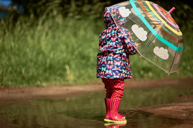 Reizend kleines Mädchen mit Regenschirm hat den Spaß, der in den gumboots im Pool nach Regen steht