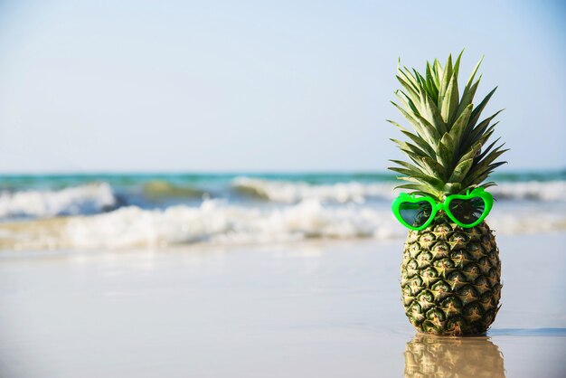 Reizende frische Ananas setzte Herzformsonnenbrillen auf sauberen Sandstrand mit Seewelle - frische Frucht mit Meersandsonnen-Ferienkonzept