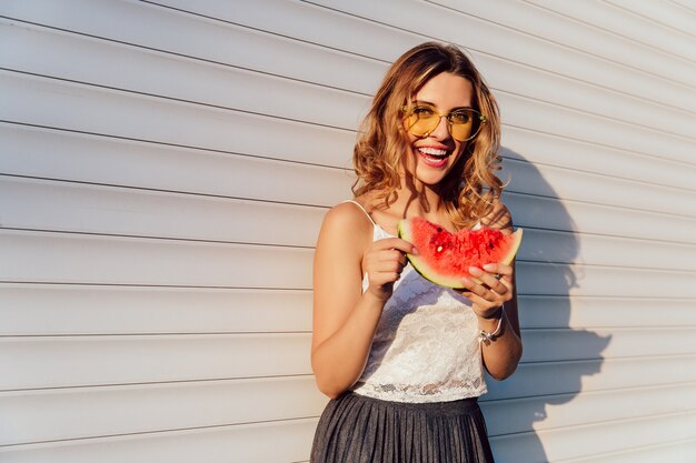 Reizendes gelocktes Mädchen hält ein Stück Wassermelone und trägt gelbe Sonnenbrille