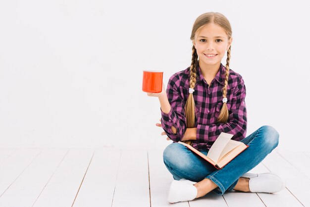 Reizendes kleines Mädchen, das ein Buch liest und einen Becher hält