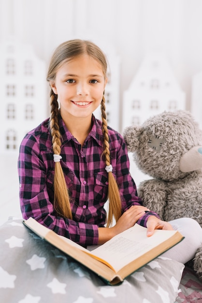 Reizendes kleines Mädchen, das ein Buch mit ihrem Teddybären liest