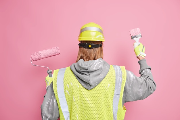 Kostenloses Foto renovierungsdienstleistungskonzept. rückansicht des rothaarigen mannes verwendet baugeräte, die in arbeitsuniform gekleidet sind und gegen rosa wand posieren. professioneller anstreicher renoviert haus