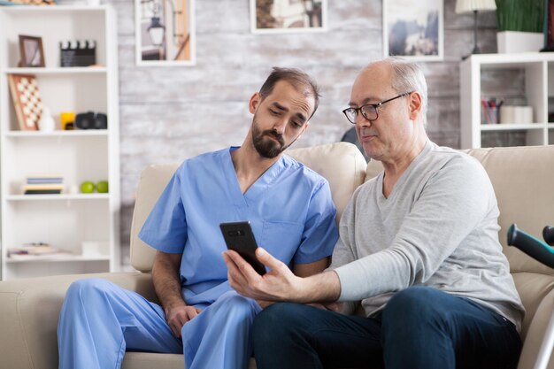 Rentner im modernen Pflegeheim sitzt mit seinem Gesundheitsbesucher auf der Couch. Älterer Mann, der Handy verwendet.