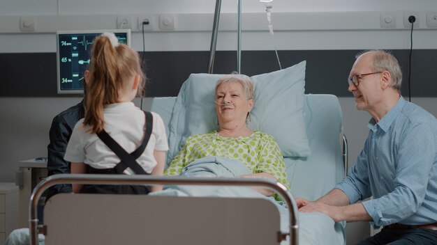 Rentnerin genießt Besuch von kleinem Mädchen und Mutter in der Krankenstation. Nichte und Tochter besuchen Oma im Krankenhaus mit Krankheit im Bett und sprechen zusammen mit Mutter und altem Mann.