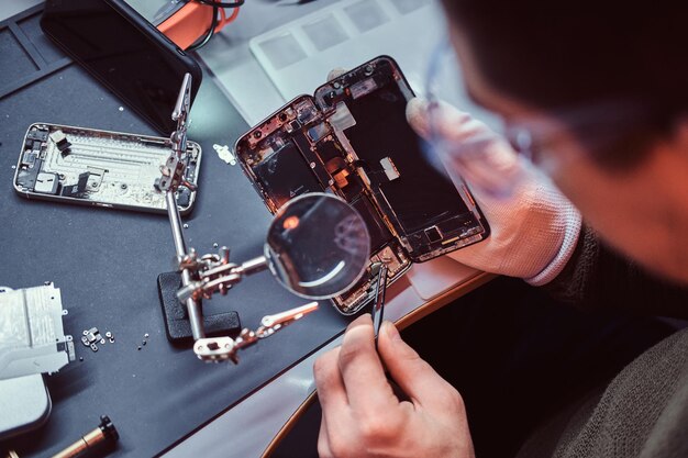 Reparateur verwendet Lupe und Pinzette, um beschädigtes Smartphone zu reparieren. Nahaufnahme eines zerlegten Smartphones.