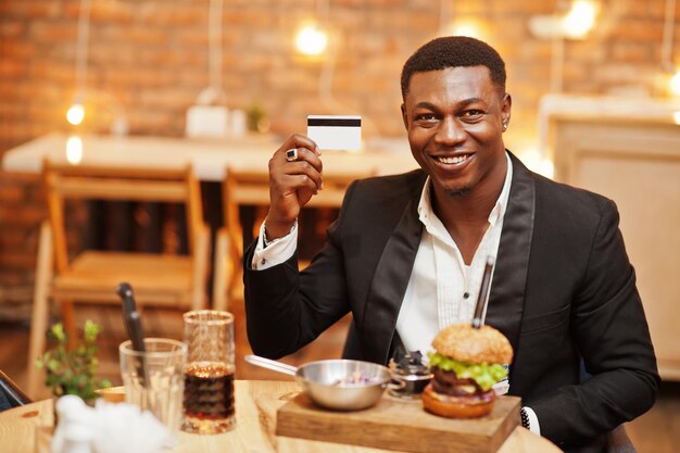 Respektabler junger Afroamerikaner im schwarzen Anzug, der im Restaurant sitzt, hält Kreditkarte mit leckerem Doppelburger und Limonadengetränk