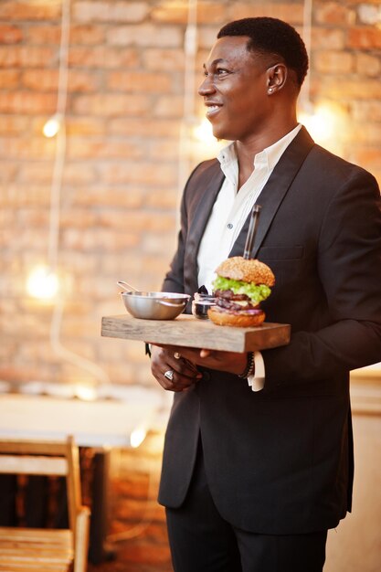 Respektabler junger Afroamerikaner im schwarzen Anzug hält ein Tablett mit Doppelburger gegen die Ziegelwand des Restaurants mit Lichtern